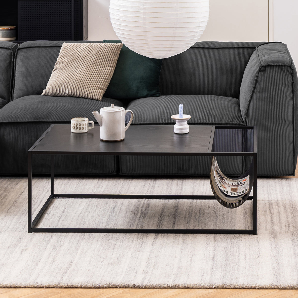 Skeleto-sofabordet (110x40) foran en grafitgrå sofa på et lyst tæppe, med en kande og magasiner i en metalpose
