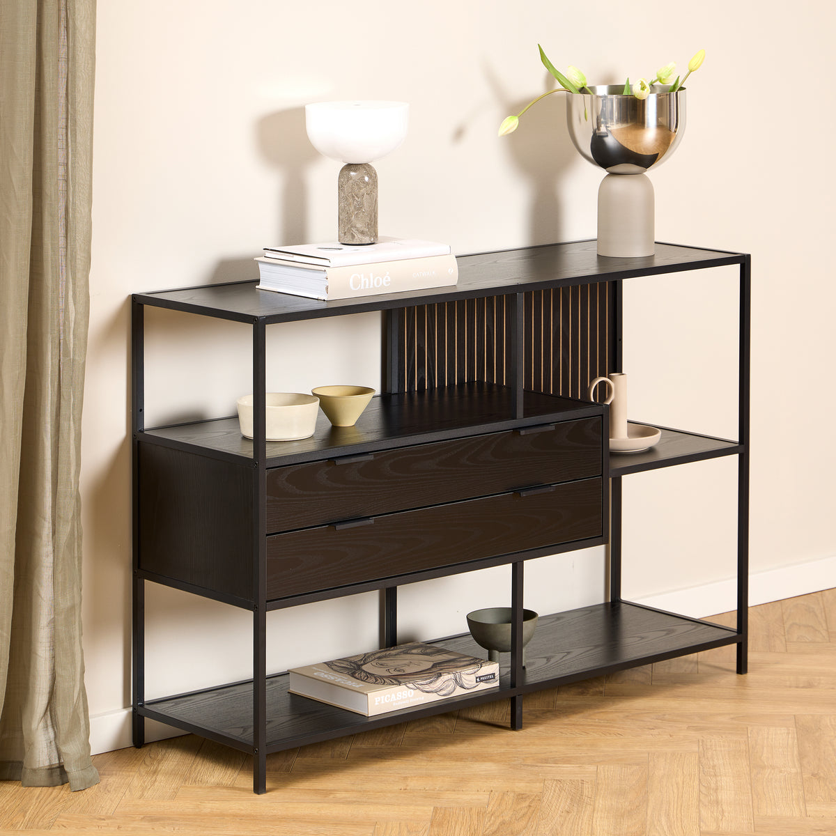 Et lavt Skeleto-reol 114x78 i sort melamin ved en lys væg, med en lampe og en vase med tulipaner på hylden