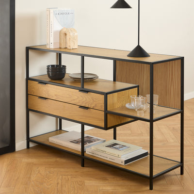 Et lavt Skeleto-reol 114x78 i farven »Vild eg« under en sort lampe, med bøger, skåle og glas på hylderne