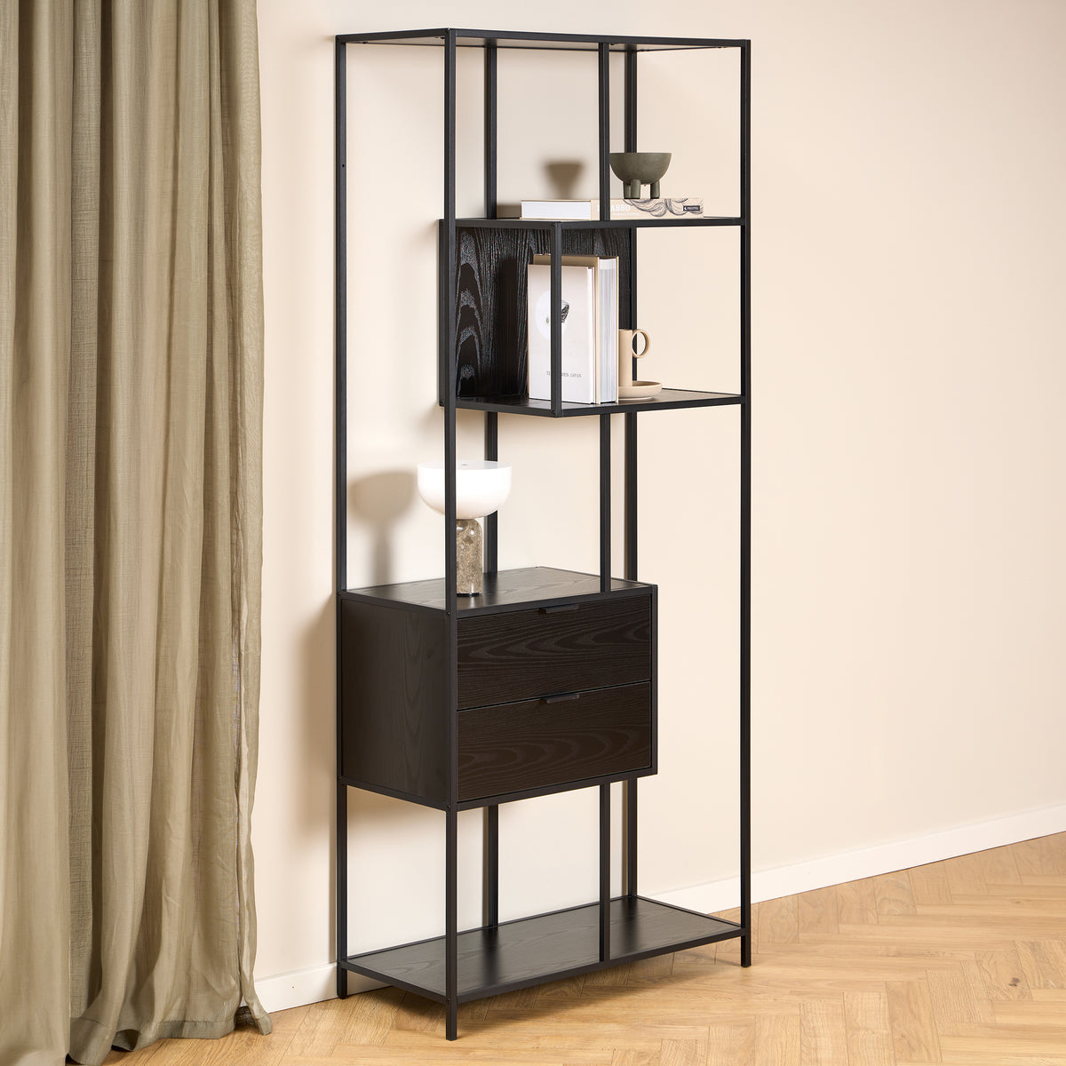 Skeleto-reol 77x185 i sort melamin ved en lys væg og et linnedgardin, med en lampe og bøger